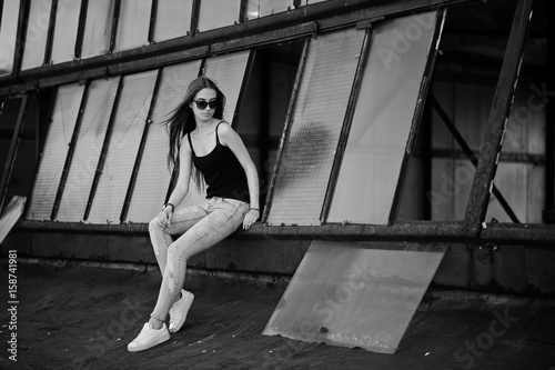 Girl at sunglasses and jeans posed at the roof of abadoned industrial place with windows.