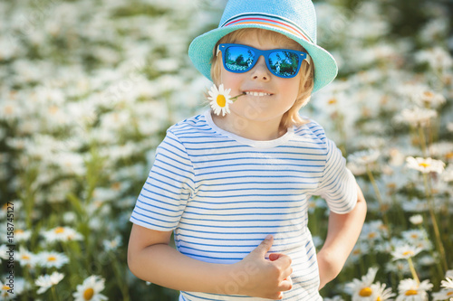 Emotional kid in the camomile field. Cheerful child with flowers wearing hat and sunglasses. Happy boy in the summer time outdoors with chamomile flower in his hand