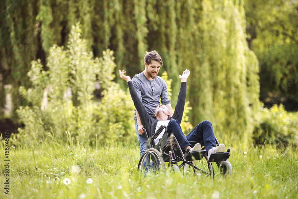 Hipster son walking with disabled father in wheelchair at park. Hipster son walking with disabled father in wheelchair at park.