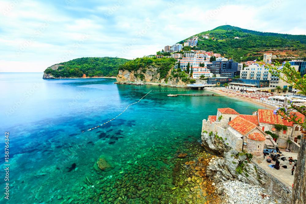 Sea landscape and beautiful cityscape. Rocky island and shoreline in cloudy evening. Old Town of Budva, medieval walled city, bay, mountains, coastal strip, sandy beaches on Adriatic Sea, Montenegro.