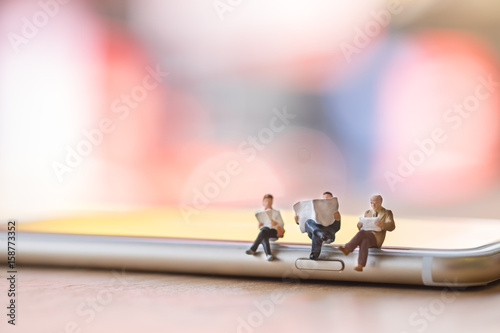 Group of miniature figures sitting and reading a book and newspaper on smart phone on wooden table. photo