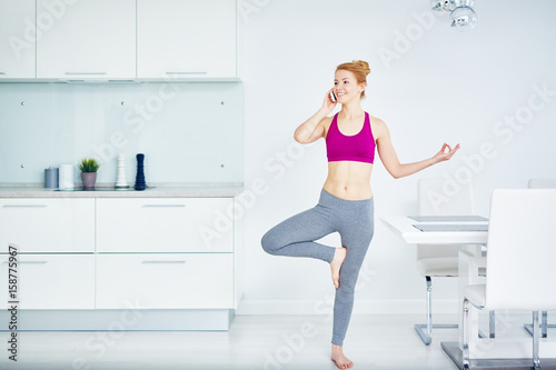 Portrait of fit red haired woman doing yoga exercises at home  standing in tree pose and speaking by phone  smiling