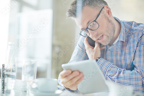 Busy middle-aged manager in eyeglasses dictating information to colleague on the phone while he using tablet photo