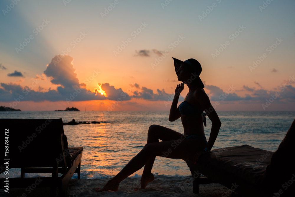 Silhouette of a beautiful slender girl on a sunset background and sun loungers on the ocean shore. Woman watch skyline on sunrise on near deck chair at the sea beach. Vacation Concept.