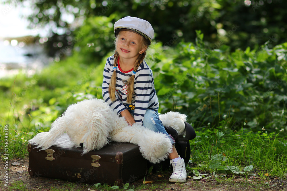 Charming cute girl sits in a summer forest on a suitcase with a soft toy in a marine striped uniform and a cap Charming cute girl sits in a summer forest on a suitcase with a soft toy in a marine striped uniform and a cap