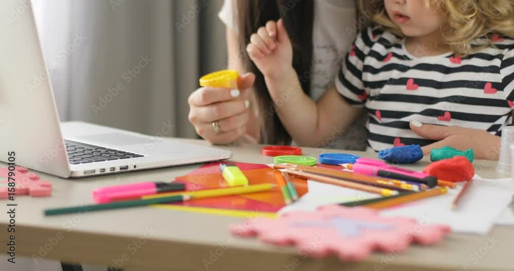 Portrait of hands of caucasian little girl with blond curly hair opening box with plasticine while her mother using laptop at home. Indoor.