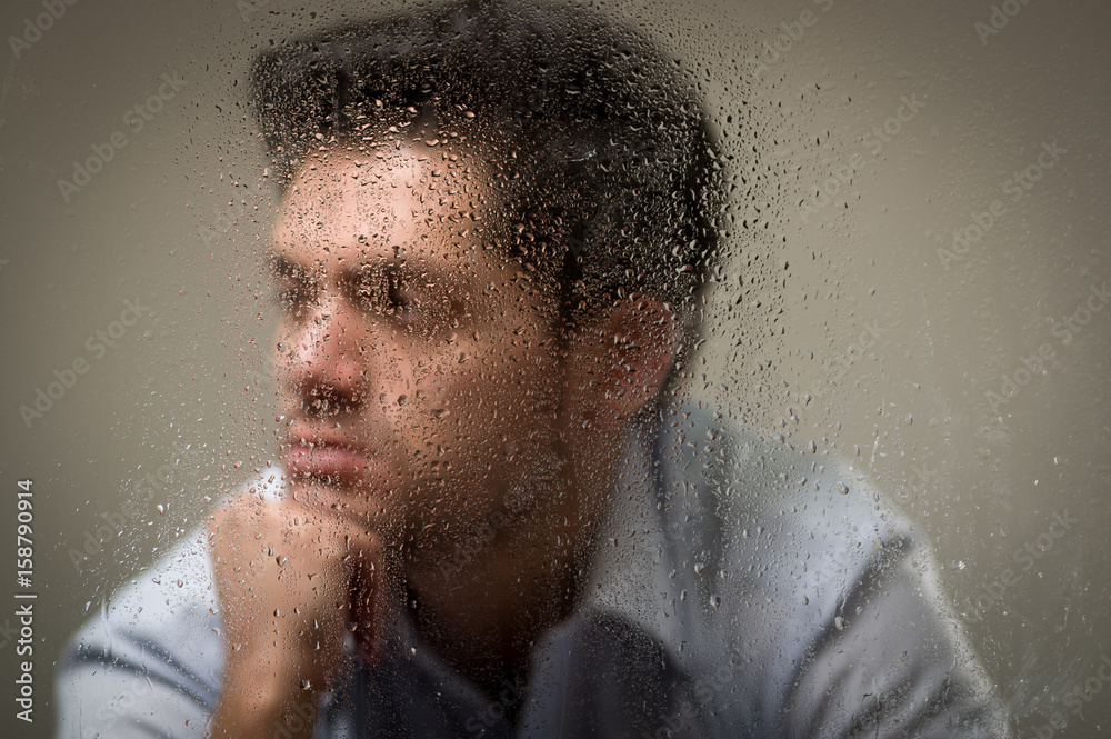 Worry depresed young man, behind a blurred window with drops, gray background