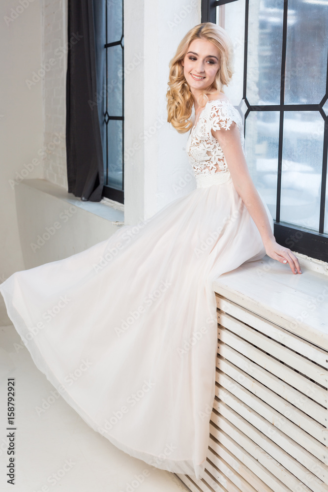 fashionable wedding gown, beautiful blonde model, bride hairstyle and makeup concept - graceful young woman in white dress sitting on the windowsill indoors, smiling female posing in the studio