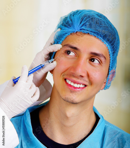 Closeup of a handsome smiling young man  receiving facial cosmetic treatment injections  doctors hand with glove holding syringe and wearing a medical hat