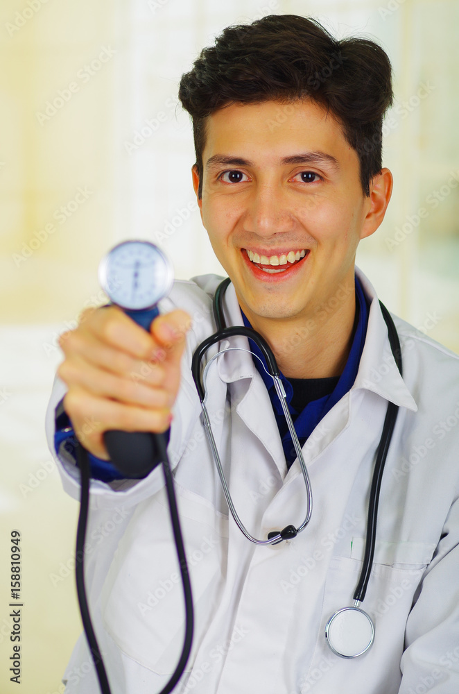 Handsome smiling young doctor with stethoscope around his neck, holdig a Tensiometer in his hand, in a doctor consulting room background Handsome smiling young doctor with stethoscope around his neck, holdig a Tensiometer in his hand, in a doctor consulting room background