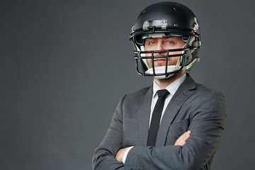 Confident businessman wearing suit and American football helmet standing cross-armed against grey background and looking at camera, copy space to left