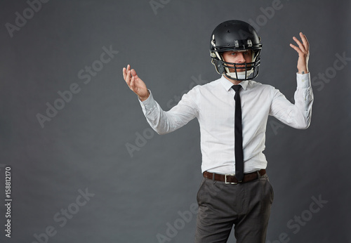Portrait of confused middle-aged businessman wearing American football helmet making hopeless gesture against grey background, copy space to the left © pressmaster