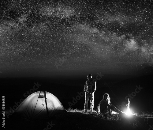 Romantic pair tourists in his camp at night near campfire and tent against starry sky. Black and white