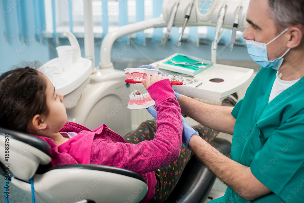 Girl in dentist chair educating about proper tooth-brushing by her pediatric dentist, using dental jaw model and toothbrush in dental office. Dentistry, early prevention, oral hygiene concept. Girl in dentist chair educating about proper tooth-brushing by her pediatric dentist, using dental jaw model and toothbrush in dental office. Dentistry, early prevention, oral hygiene concept.