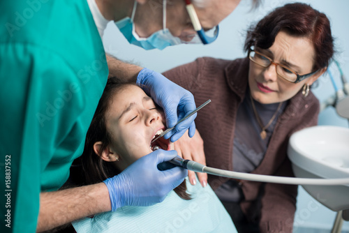 Girl with her mother on the first dental visit. Senior male dentist doing dental procedures of patient at the dental office. Dentistry  medicine  stomatology and health care concept. Dental equipment