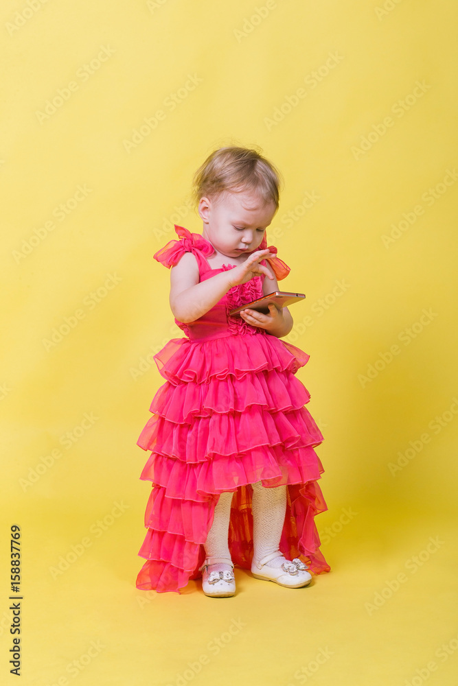 Girl toddler in a pink dress on a yellow background holding a smartphone