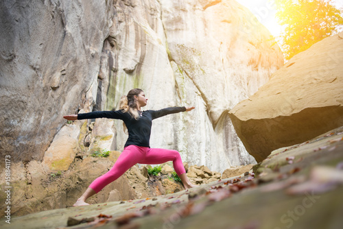 Sporty slim woman in pink footless is practicing yoga and doing asana Virabhadrasana 2 outdoors. Huge rocks on the background photo