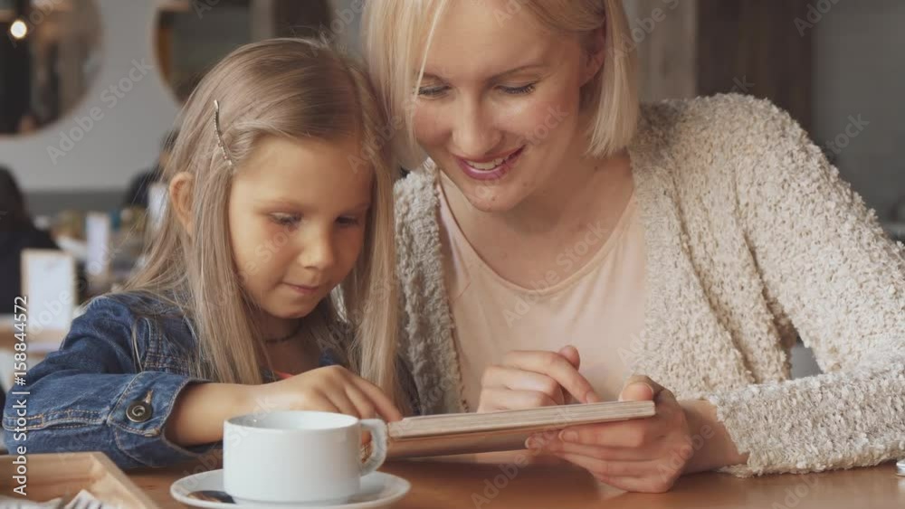 Attractive blond mother and her daughter choosing drinks at the cafe. Close up of pretty young woman holding menu in her hand. Cute little girl showing her mother what she wants to drink Attractive blond mother and her daughter choosing drinks at the cafe. Close up of pretty young woman holding menu in her hand. Cute little girl showing her mother what she wants to drink