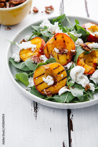 Fresh salad with grilled peach halves, arugula and burrata on a plate on white distressed wooden background. Top view. Summer food concept