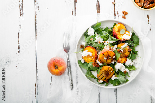 Fresh salad with grilled peach halves, arugula and burrata on a plate on white distressed wooden background. Top view, horizontal composition with copy space. Summer food concept