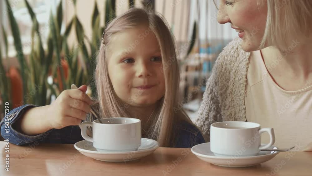 Cute little girl moving her teaspoon in the cup at the cafe. Close up of pretty blond mother and her daughter chatting at the table. Attractive caucasian female child nodding to her mom