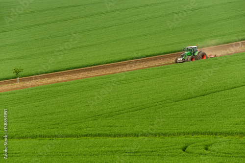 Obraz na płótnie Scenic View Of Modern Farming Tractor Which Plowing Green Field. Agriculture Tractor Cultivating Wheat Field And Creating Green Abstract Background Texture.Big tractor working on colorful spring field w salonie