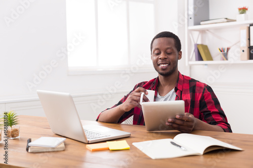Black businessman in casual office, reading news on tablet, drinking coffee