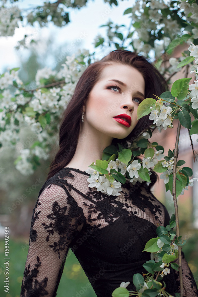 Fototapeta premium Fashion art Woman in summer near blossoming Apple tree, bright lipstick. Spring summer mood, mysterious romantic image of girl in a black dress tree flowers. Woman dreams