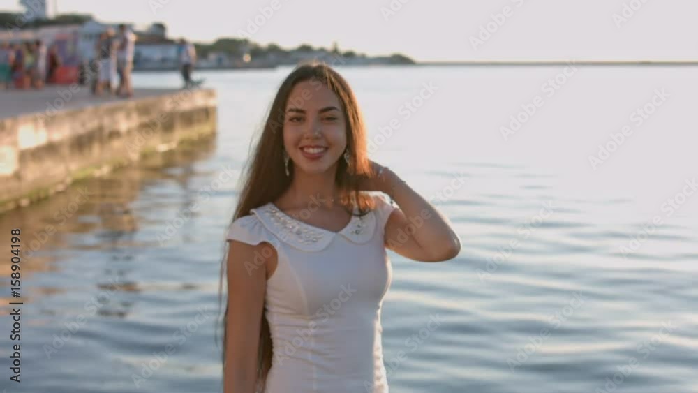 Stunning brunette with long hair dressed in the white tight dress standing by the sea Stunning brunette with long hair dressed in the white tight dress standing by the sea
