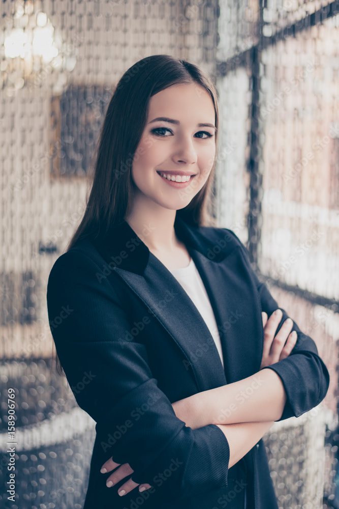 Close up cropped portrait of successful and confident business lady standing with crossed hands at her work station in modern coworking office, looks in the camera, She is in a black jacket, smiling