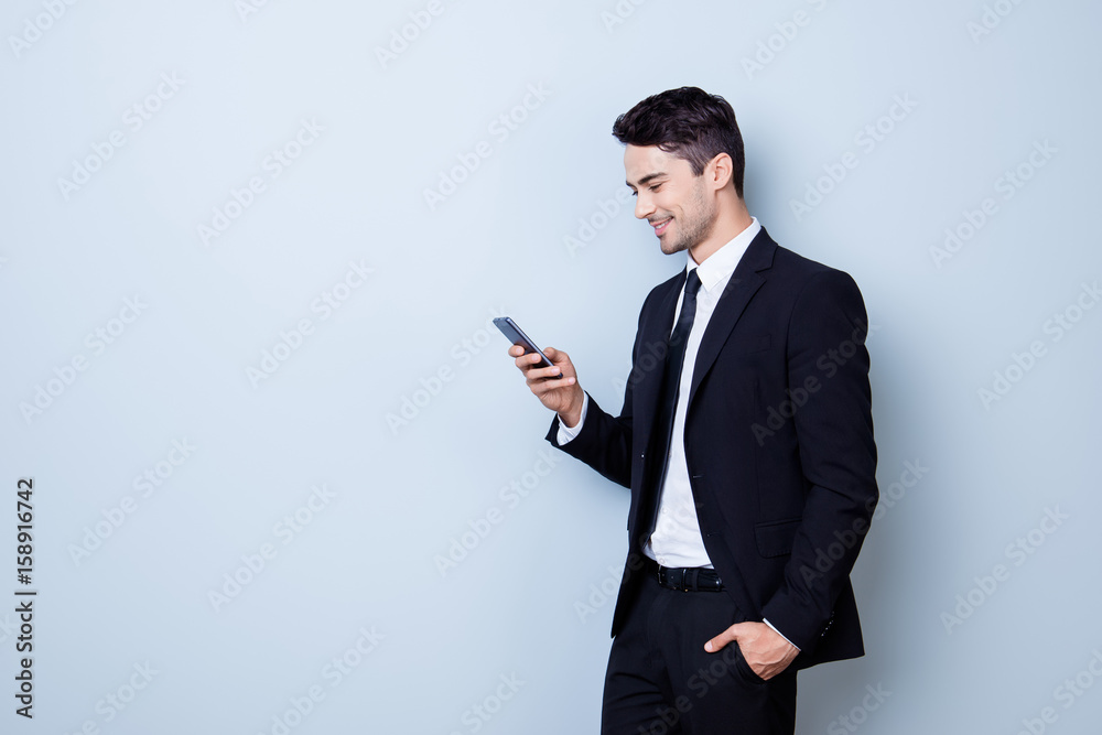 Young cheerful brunete lawyer is standing on the pure light blue background and smiling, reading good news on internet at his mobile phone, wearing suit and tie Young cheerful brunete lawyer is standing on the pure light blue background and smiling, reading good news on internet at his mobile phone, wearing suit and tie
