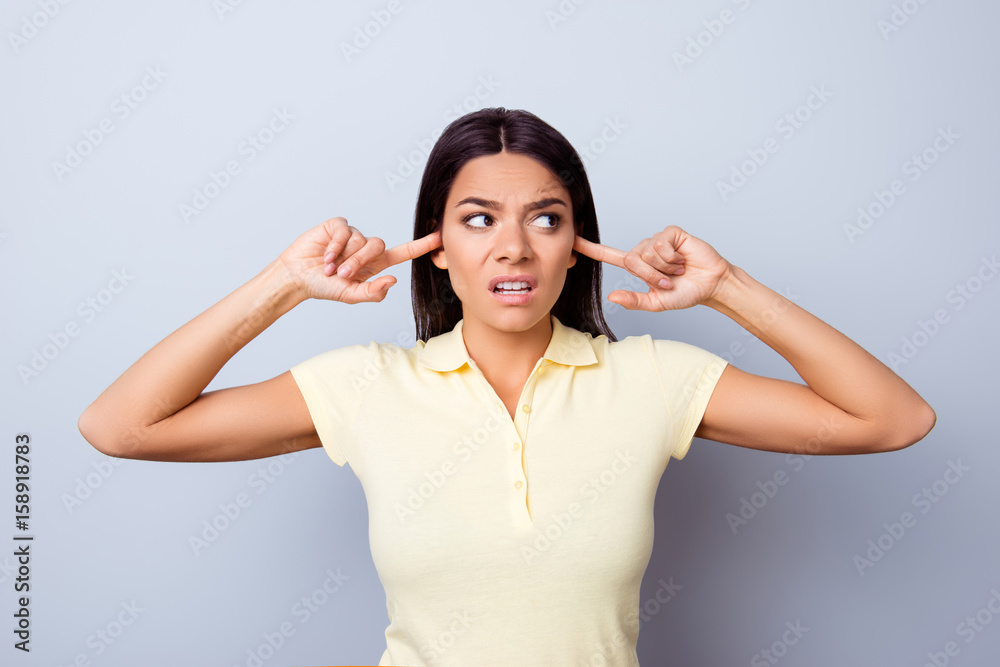 Close up of annnoyed young latinamerican girl, closing her ears with fingers. She is lookiing away  angry, she is in a casual tshirt on pure background