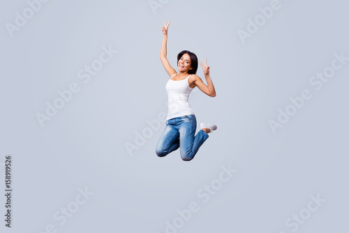 Go crazy! Happy young brunette latin girl is jumping and fooling around. She is in casual outfit, showing v sign, isolated on a pure light background