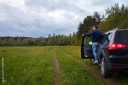 The man standing on the running Board of the jeep with the door open and looks into the distance. Traveler stands on step of all-terrain 4x4 vehicle before board barn. photo