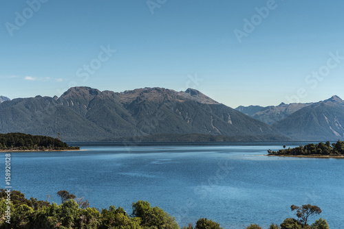 Te Anau, New Zealand - March 16, 2017: Looking over part of majestic lake Te Anau with dark blue water, under light blue sky. Big mountainous slopes with forests and other green vegetation foreground.