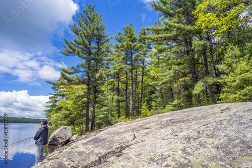 Tall pine forest towering over Canadian shield bedrock beside blue lake where man stands fishing