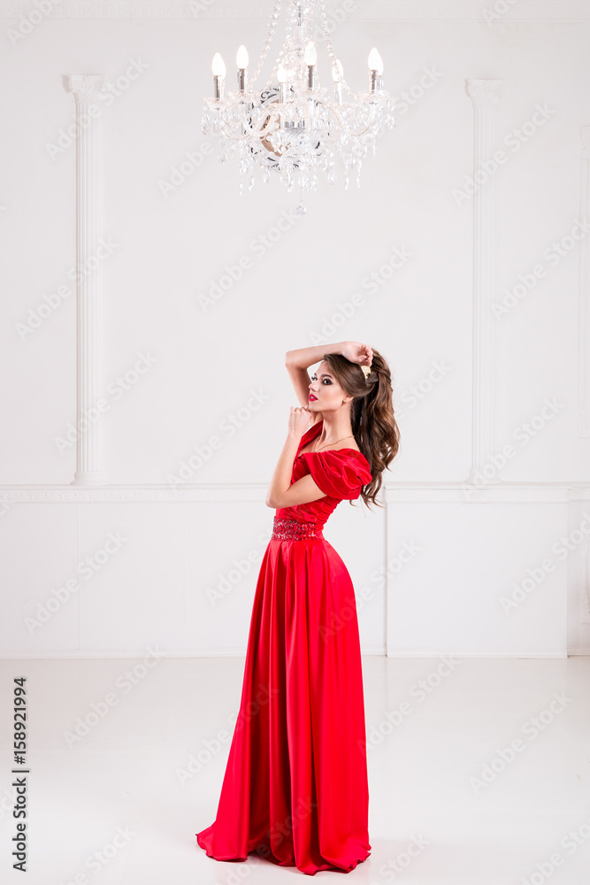 Elegant woman in a long red dress is standing in a bright room with a sofa and a vintage chandelier Elegant woman in a long red dress is standing in a bright room with a sofa and a vintage chandelier