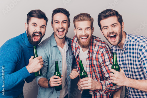 Men`s club. Four crazy friends guys are screaming with bottles in hands, all in casual shirts, isolated on grey background. They are fans of sports games as football, basketball, hockey, baseball photo