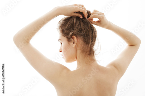 Portrait of young beautiful tender naked girl correcting bun standing back to camera over white background.