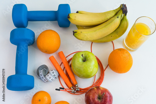 healthy eating concept. Dumbbells, skipping rope, bananas, oranges, measuring tape waist, oranges, red apple, green apple, orange juice on a white table. Fitness diet. view from above