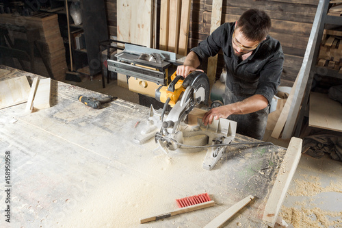 A young male carpenter builder saws a modern circular saw a wooden board in the workshop room