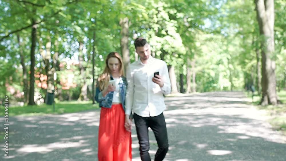 Happy couple holding hands and using smartphones while walking in the park, steadycam shot
Happy couple holding hands and using smartphones while walking in the park, steadycam shot