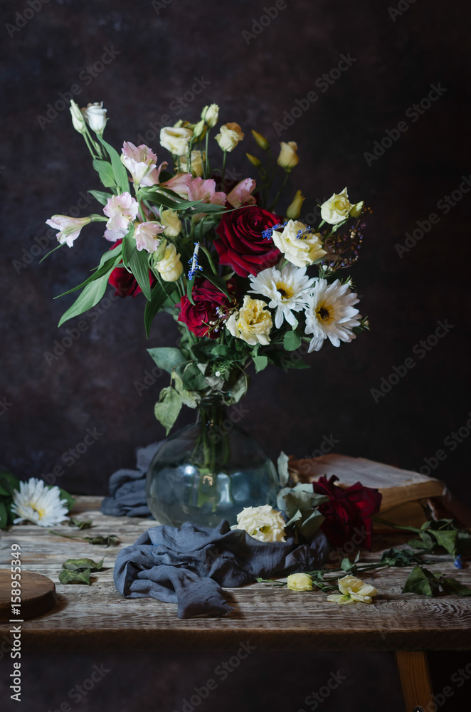vase with a bouquet of assorted flowers on a wooden table with old books, grey cloth, petals on a brown background vase with a bouquet of assorted flowers on a wooden table with old books, grey cloth, petals on a brown background