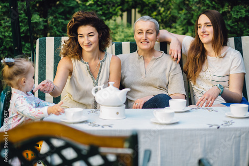 Tableau sur toile Family of four women - granddaughter with her mother, grandmother and aunt sitting and drinking tea at hte garden in vacation