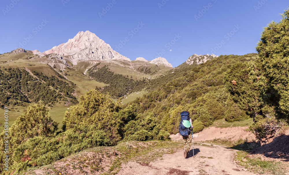 Mountain landscape. Male tourist walking on the road in the mountains, carries a heavy backpack. In the background, mountains, blue sky
