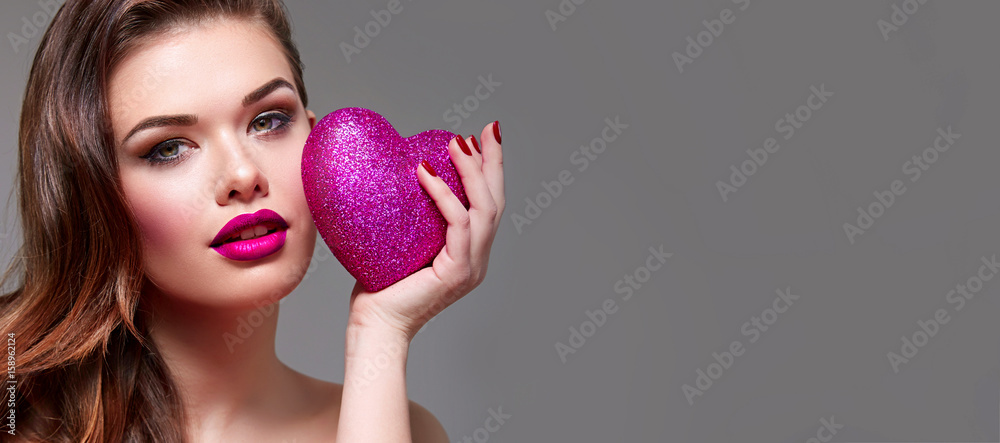 Fototapeta premium Beautiful young attractive girl in studio on a gray background holds in hands a large shiny pink heart. Romance, love. Makeup - pink lips, golden shadows. Hairstyle is a Hollywood wave.