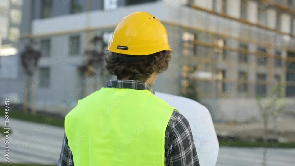 Back view of architect in yellow helmet and green vest standing on building background, analysing drawing. Outdoor. Back view of architect in yellow helmet and green vest standing on building background, analysing drawing. Outdoor.