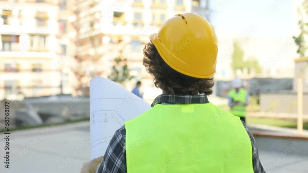 Back and side view of caucasian builder in yellow helmet and green vest looking drawing standing on unfinished construction background. Outdoor.