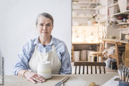 Portrait of confident senior female potter sitting at table with vase against white wall in workshop photo