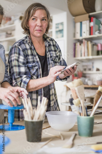 Mature female potter using mobile phone by senior colleague molding clay at workbench in store photo